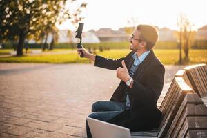 Businessman vlogger in coat wearing eyeglasses and showing thumb up while using mobile phone with gimbal. He is sitting on a bench in the park and recording vlog on a sunny autumn afternoon. photo