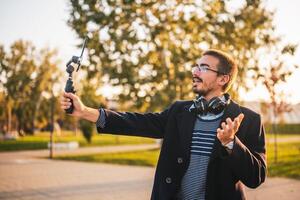 Businessman vlogger in coat wearing eyeglasses and headphones. He is using mobile phone with gimbal in the park and talking while recording vlog on a sunny autumn afternoon. photo