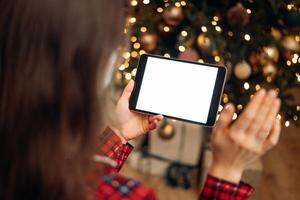 Woman holding tablet with blank screen in front of Christmas tree, festive bokeh lights in background, ready for holiday call or virtual greeting. photo