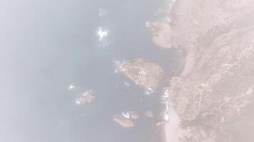 Aerial view descending through fog towards a rocky coastline with visible rugged terrain and ocean waves breaking near the shore video