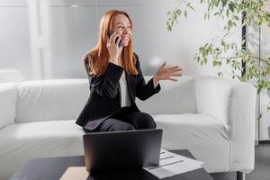 Smiling middle-aged female employee wearing glasses makes a call at work while sitting at her desk and talking to a client or customer on the webcam. photo