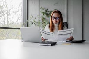 Senior chic woman using laptop at home while taking notes in notebook. Old freelancer works in the living room on a laptop while drafting details on a book. photo
