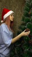 Vertical photo of girl in New Year cap decorating artificial New Year tree with pine cone. Folding Christmas tree for home decoration for New Year and Christmas