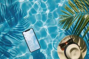 Mobile phone mockup with blank screen laying on the blue water of the sea or pool with palm tree, sunglasses and hat photo