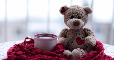 Valentines Day still life with steaming mug of tea and stuffed toy teddy bear with red heart shaped patch. Cup and bear wrapped in red crochet scarf on table under snowfall. Love greeting 4K video