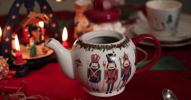 Hand put red cap atop Christmas teapot with nutcrackers pattern closeup. Pouring hot water in crockery teapot. Christmas table setting with candle and nativity carousel. Steam coming out from kettle video