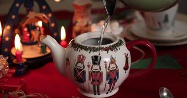 Christmas teapot with nutcrackers pattern and red cap closeup. Pouring hot water in crockery teapot. Christmas table setting with candle and nativity carousel. Steam coming out from kettle. New Year video