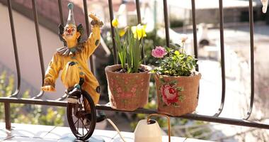 Brightly colored burlap flower pots are displayed on a balcony railing, featuring blooming flowers. A cheerful clown figurine enhances the lively atmosphere of this charming outdoor space. video