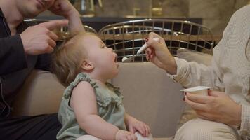 Little girl enjoys eating yogurt with a spoon that her mother is feeding her. The baby's face is sweetly covered in yogurt. video