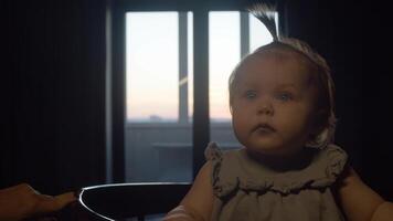 Little one-year-old girl is making faces in her crib in evening, against backdrop of sunset that can be seen from apartment window, before going to bed. video