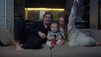 Parents are laughing while watching TV, sitting on couch with their baby. They are actively pointing to screen and showing their child what they are watching. video