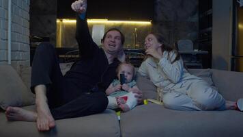 Joyful family spends quality time together, watching TV and relaxing on couch. Father decides to have some fun with his wife and child, trying to get them laughing by making silly faces and imitating characters from cartoon. video