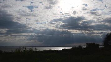 Sunbeams break through grey cumulus clouds in sky before rain. River shore landscape with silhouettes of plants swaying in wind. Cloudscape at countryside garden with shiny lake water on background video