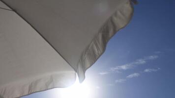 Summer sunshine in blue sky framed by frills of white beach umbrella flutter on breeze. View from under beach sunshade on sky background with copy space. Protection from overheating on beach in summer video