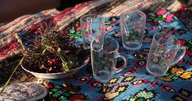 Herbal ingredients are arranged neatly alongside four transparent cups on a vibrant textile. The sun casts warm light, enhancing the natural colors of the setup. video