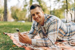 Portrait of attractive, smiling man holding, using smartphone, lying on blanket, relaxing photo