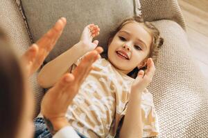 Happy, smiling little daughter playing clapping hands with mother lying on comfortable sofa photo