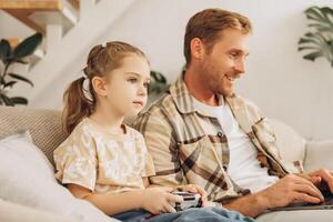 Little daughter holding console playing game, father using laptop, typing on keyboard photo