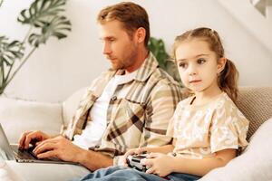 Little daughter holding console playing game, father using laptop, typing on keyboard photo