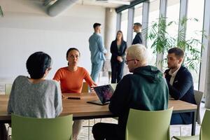 A diverse team of business professionals engaged in a discussion around a conference table in an office, while their colleagues collaborate in the background photo