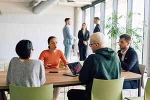 A diverse team of business professionals engaged in a discussion around a conference table in an office, while their colleagues collaborate in the background photo