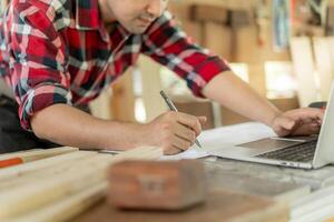 Asian man owner a small furniture business is recheck order online. carpenter male is adjust wood to the desired size. architect, designer, Built-in, professional wood, craftsman, workshop. photo