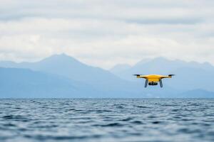 A yellow drone flying over a calm ocean with mountains in the background on a cloudy day. photo
