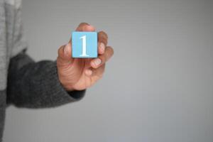 A hand is holding a blue block that has the number one inscribed on it against a neutral background photo