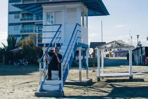 young man with a backpack sits on the steps of a lifeguard tower near the sea photo