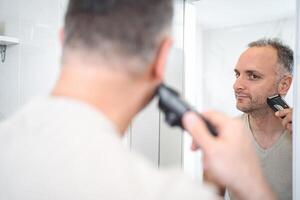 Man shaves his beard with a trimmer in the bathroom photo