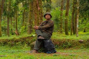 An adventurer is sitting on a large rock in a dense pine forest. An Asian man in a flannel shirt and bucket hat is sitting and resting while enjoying the tranquility of the beautiful nature. photo