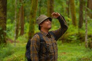 Half-body close-up of an adventurer in a dense and green pine forest. An Asian male traveler in a flannel shirt, bucket hat and backpack poses against a backdrop of dense pine trees. photo