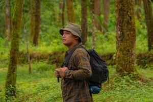 Half-body close-up of an adventurer in a dense and green pine forest. An Asian male traveler in a flannel shirt, bucket hat and backpack poses against a backdrop of dense pine trees. photo
