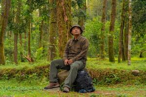 An adventurer is sitting on a large rock in a dense pine forest. An Asian man in a flannel shirt and bucket hat is sitting and resting while enjoying the tranquility of the beautiful nature. photo
