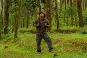 Asian man in adventure outfit in dense and green pine forest area. An adventurer in a flannel shirt, bucket hat, and backpack poses against a backdrop of dense pine trees. photo