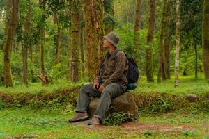 An adventurer is sitting on a large rock in a dense pine forest. An Asian man in a flannel shirt and bucket hat is sitting and resting while enjoying the tranquility of the beautiful nature. photo