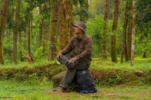 An adventurer is sitting on a large rock in a dense pine forest. An Asian man in a flannel shirt and bucket hat is sitting and resting while enjoying the tranquility of the beautiful nature. photo