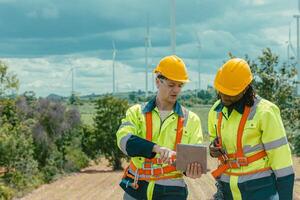 Engineer workers using tablet computer service monitor Wind Turbines status, working survey at wind electricity generator farm field outdoor. photo