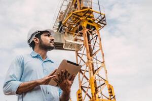 Smart Indian worker engineer manager manage construction site. Architect foreman with tower crane and tablet computer. photo