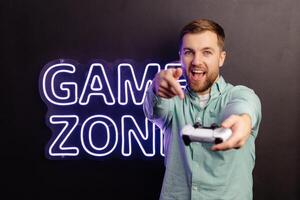 Young bearded man standing at the wall in the room and playing, holding wireless joystick, screaming, and winning intense game match photo