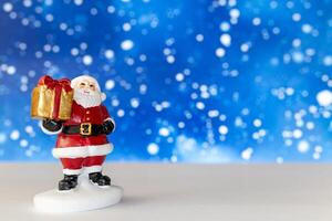 Cheerful Santa Claus figurine holding a golden gift box, set against a vibrant blue background with snowy bokeh effects photo