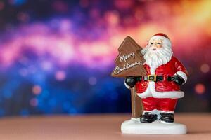 Santa Claus figurine holding a Merry Christmas tree sign, set against a glowing bokeh background in warm festive tones photo