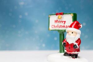 Miniature Santa Claus figurine standing next to a festive Merry Christmas sign photo