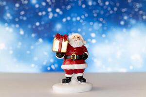 Cheerful Santa Claus figurine holding a golden gift box, set against a vibrant blue background with snowy bokeh effects photo