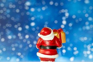 Cheerful Santa Claus figurine holding a golden gift box, set against a vibrant blue background with snowy bokeh effects photo