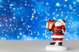Cheerful Santa Claus figurine holding a golden gift box, set against a vibrant blue background with snowy bokeh effects photo
