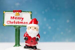 Miniature Santa Claus figurine standing next to a festive Merry Christmas sign photo