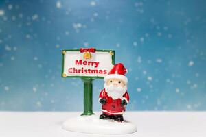 Miniature Santa Claus figurine standing next to a festive Merry Christmas sign photo