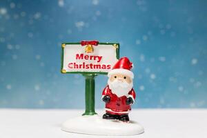 Miniature Santa Claus figurine standing next to a festive Merry Christmas sign photo