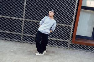 A young man in a grey sweatshirt and black pants poses in front of a chain link fence photo